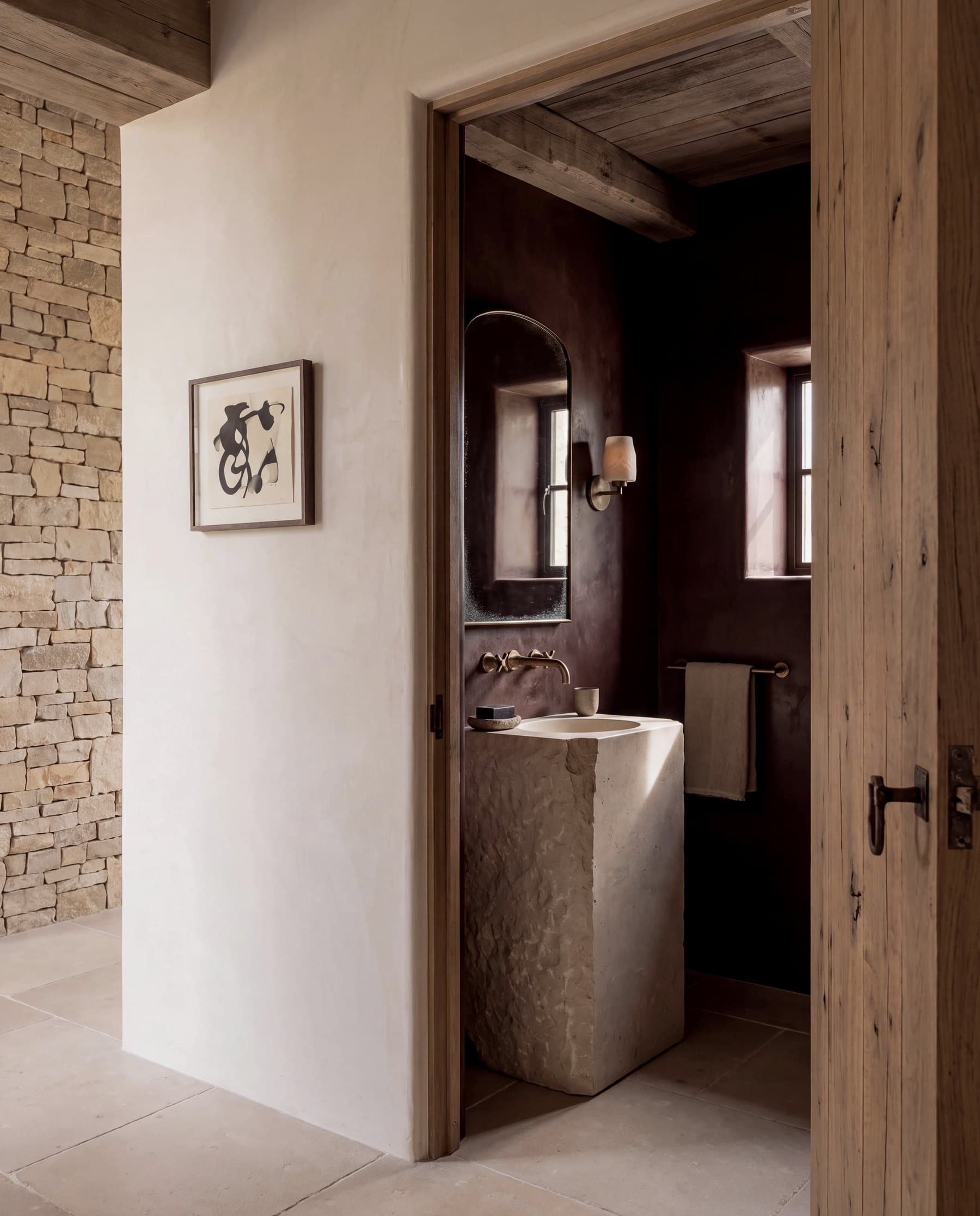 Powder room in deep oxblood plaster with a rough-hewn limestone pedestal basin, seen through an open oak door