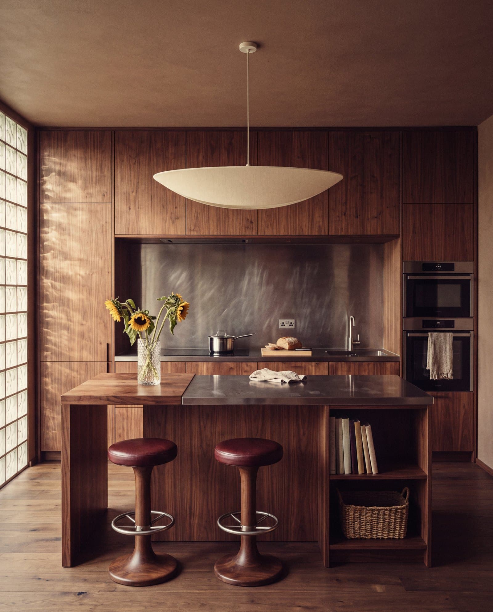 Walnut kitchen with a brushed steel backsplash, glass-block window and two leather stools at the island